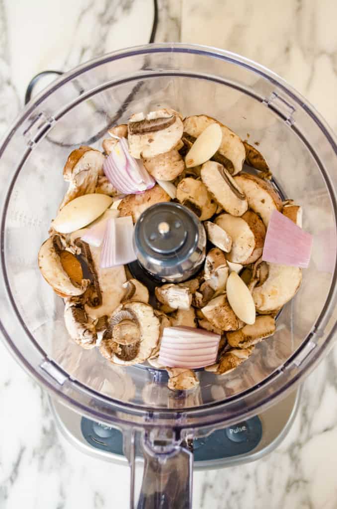 Looking down into a food processor containing sliced brown mushrooms, chunks of shallot and a few cloves of garlic to make Easy Meatball Wellingtons - The Goldilocks Kitchen