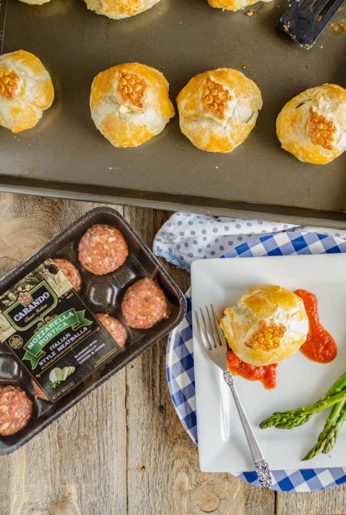 An Easy Meatball Wellington on a plate next to a package of Carando Meatballs. Adjacent is a rimmed baking sheet with more Easy Meatball Wellingtons. - The Goldilocks Kitchen