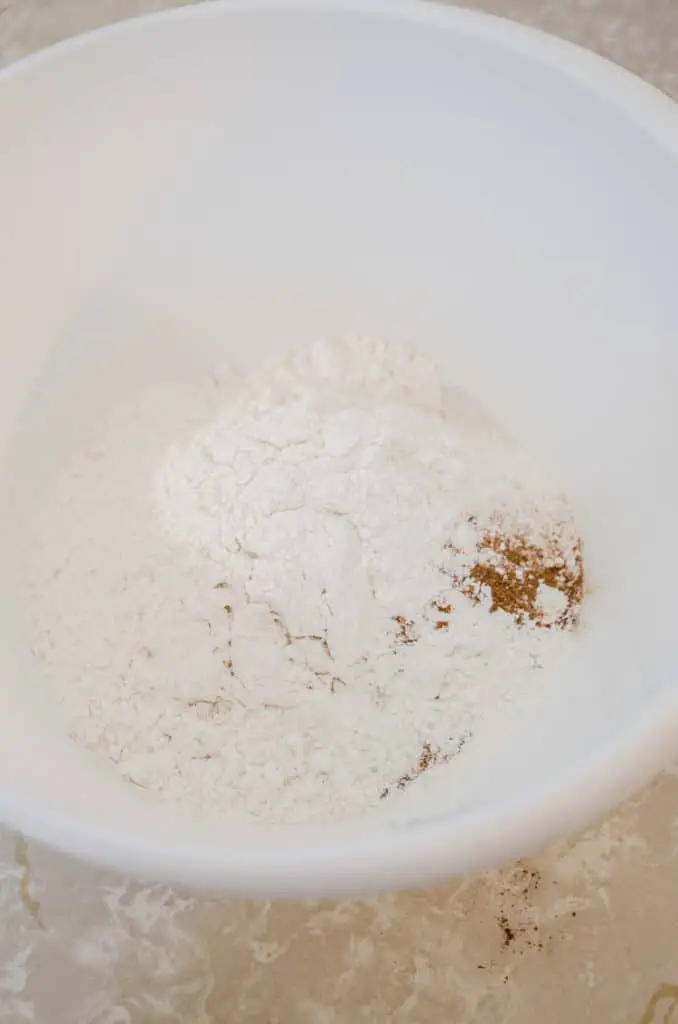A white mixing bowl contains white flour, cinnamon, allspice, salt, and baking soda for making Apple Cinnamon Cupcakes - The Goldilocks Kitchen