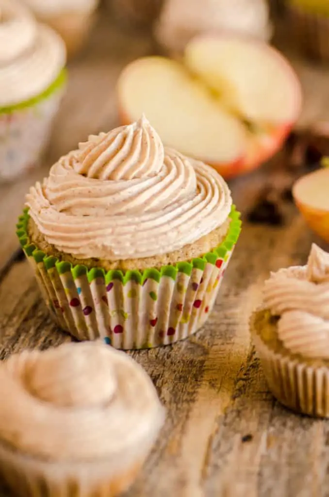 Apple Cinnamon Cupcakes with cinnamon buttercream frosting swirled on top are displayed on a wooden table with apple slice in the background- The Goldilocks Kitchen