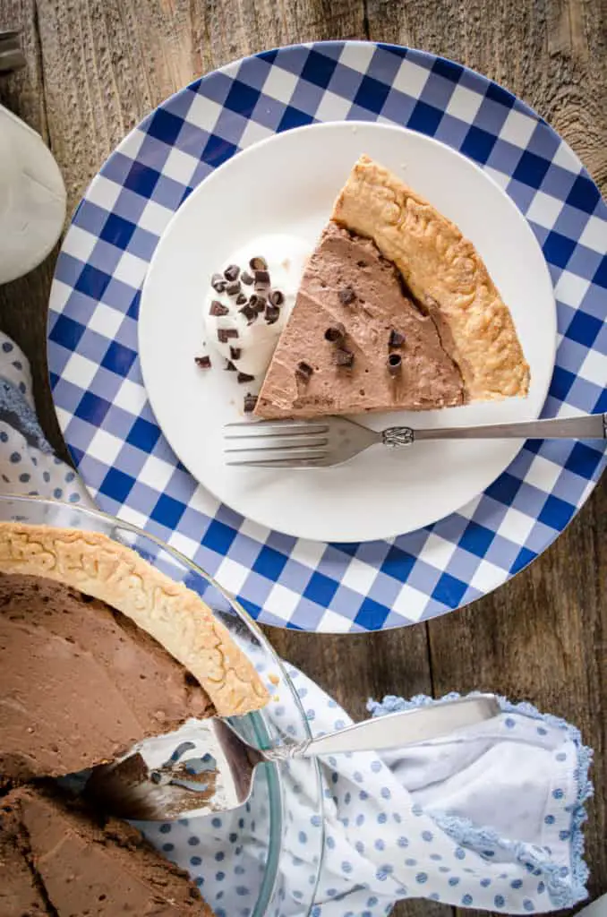 A slice of French Silk Pie without Raw Eggs sits on a blue and white gingham plate garnished with chocolate curls- The Goldilocks Kitchen