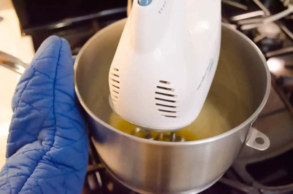 A hand held mixer beats egg whites in a metal bowl set over a pot of simmering water to cook the egg whites to make French Silk Pie without Raw Eggs - The Goldilocks Kitchen