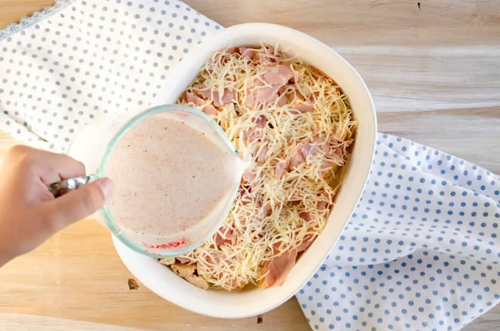 Assembling an Easy Weeknight Chicken Cordon bleu Casserole with a seasoned white sauce being poured over the top.