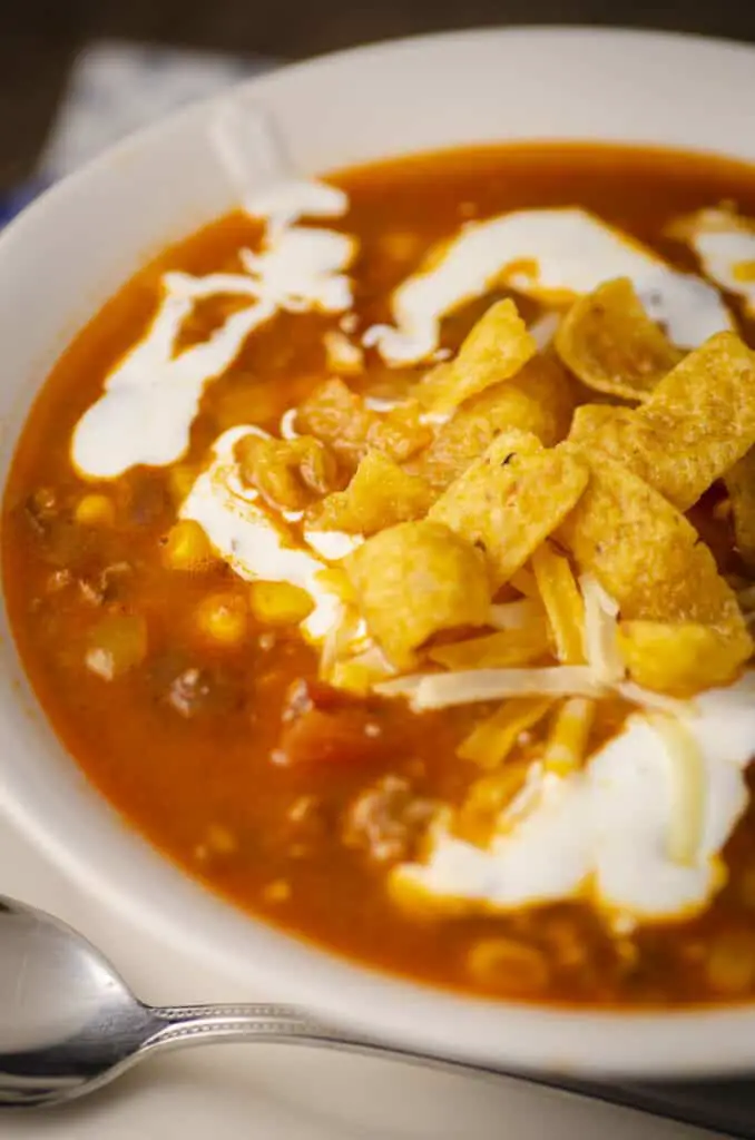 An extreme closeup of a bowl of Weeknight Taco soup showing corn, beans, tomatoes and ground beef in broth. The bowl is garnished with sour cream, shredded cheese and tortilla strips.