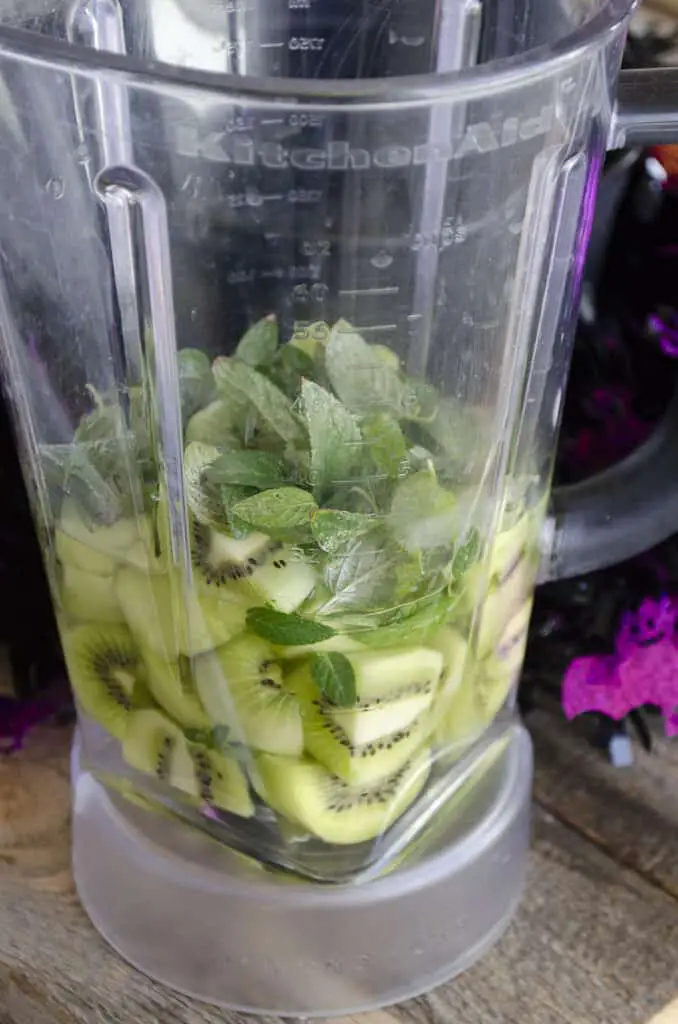 A blender pitcher sits on a wooden table filled with sliced green kiwifruit, mint leaves and clear simple syrup to make Witches Brew with Dry Ice.
