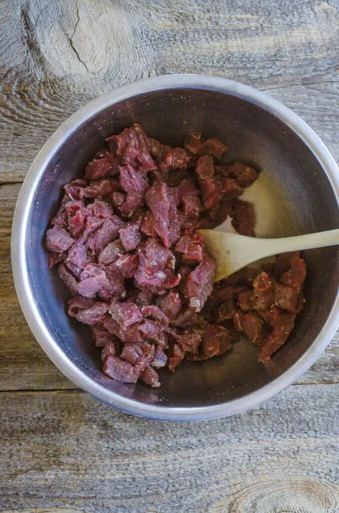 Bite sized pieces of raw beef are stirred together with seasonings in a bowl.