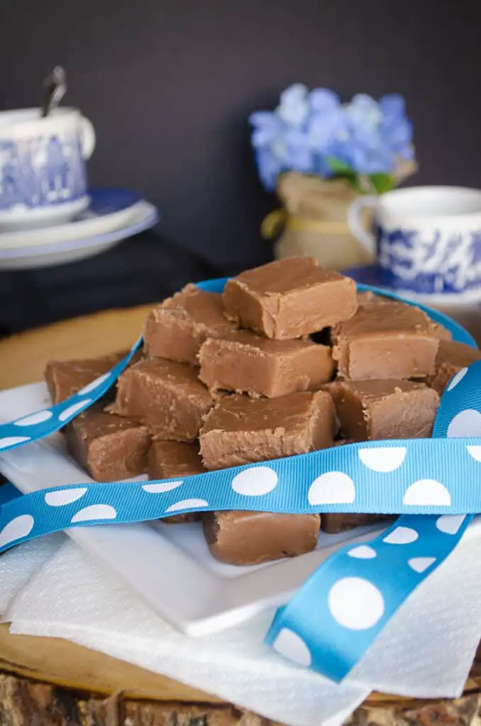 Blue-Ribbon Fudge squares sit on a white square plate with China tea cups and blue flowers in the background.