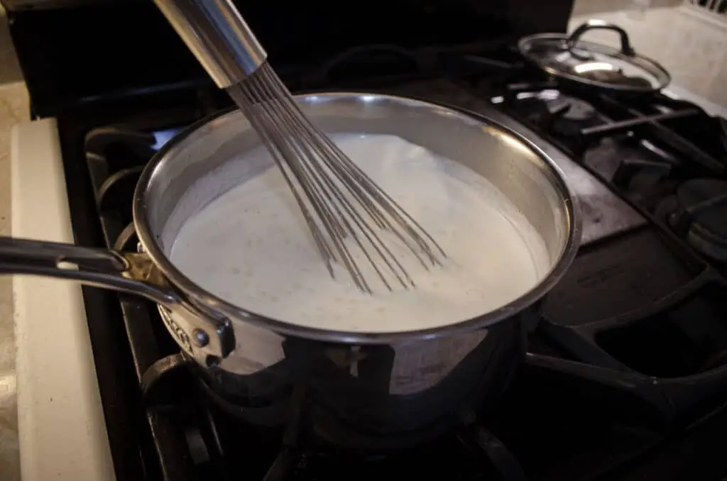 A whisk stirs hot milk and tapioca pearls in a pot on a gas stove. 