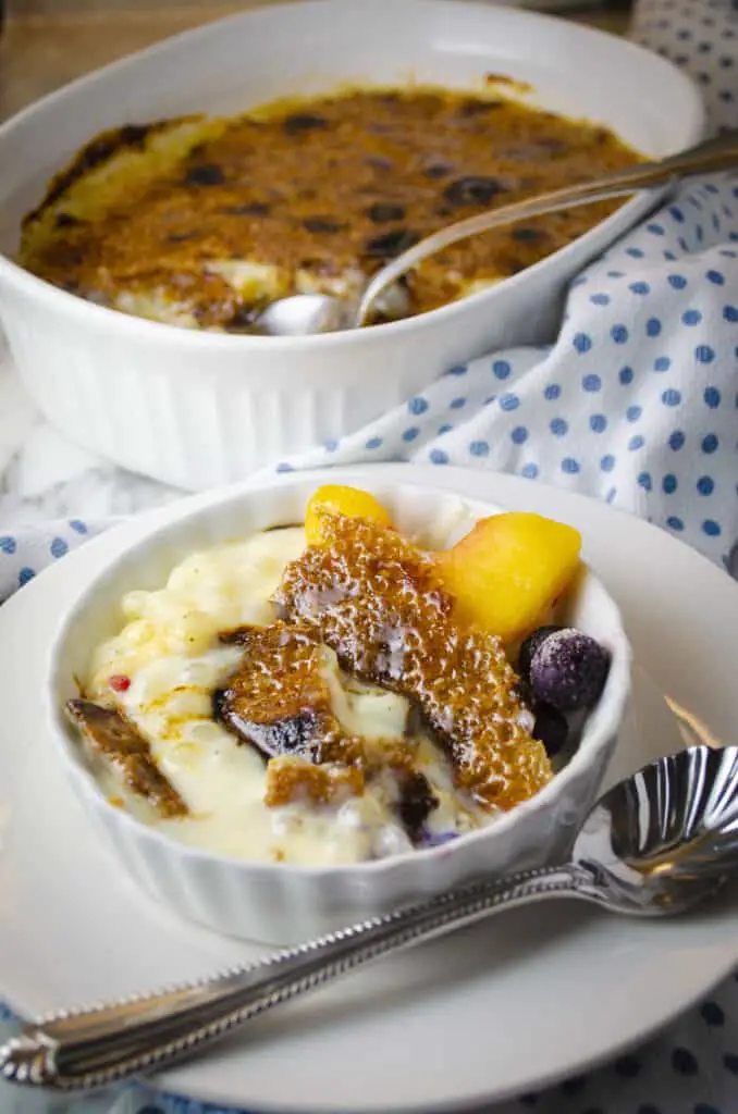 A white ceramic serving dish with Tapioca Pudding Brûlée in it, garnished with a peach slice and some blueberries. A Large baking dish with Tapioca Pudding Brûlée sits behind it in the background.