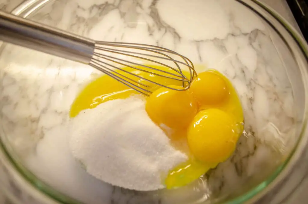 A clear glass bowl containing three egg yolks and white sugar.