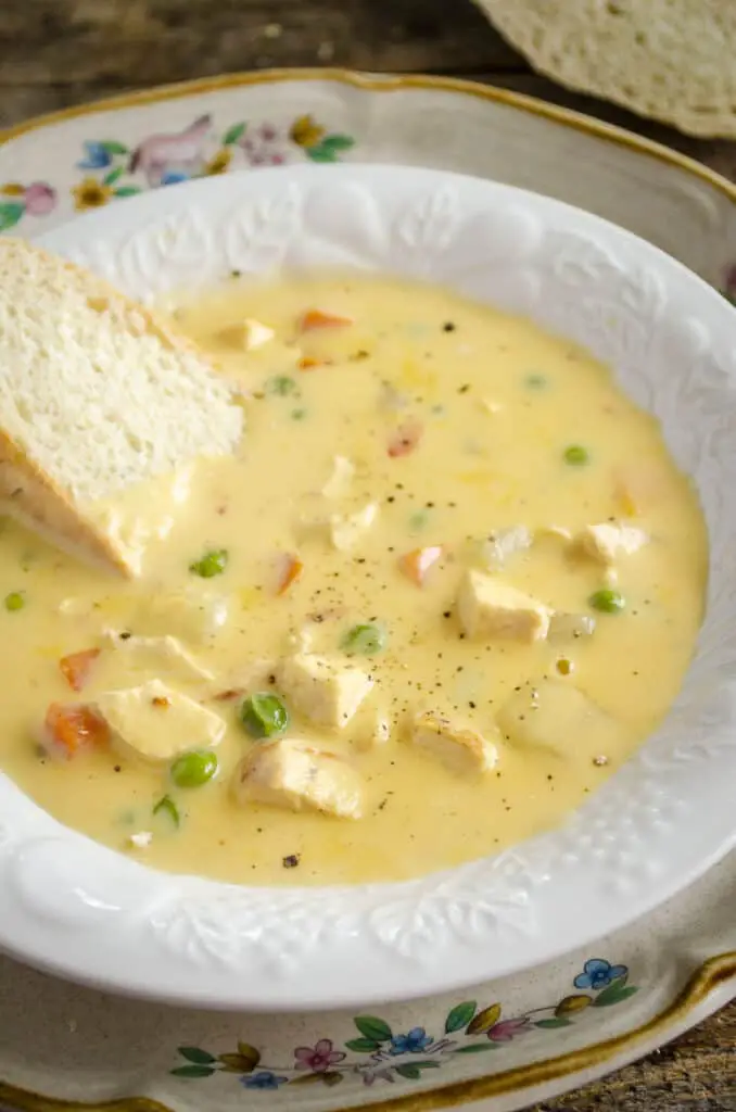 A white decorative ceramic bowl is filled with 20-Minute Cheesy Chicken Soup with a slice of crusty bread dunked into one side of the bowl.