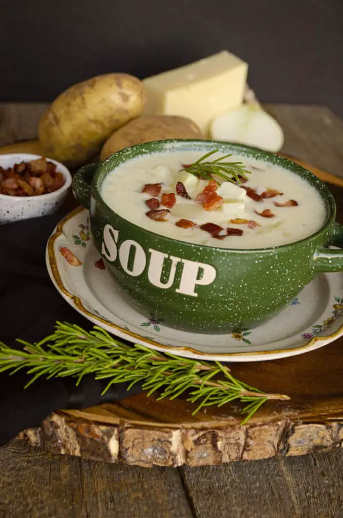 Looing at a green bowl with the word "soup" on it's side, filled with Gluten-free Swiss Potato Soup and surrounded by ingredients such as swiss cheese, raw potatoes and onion. 