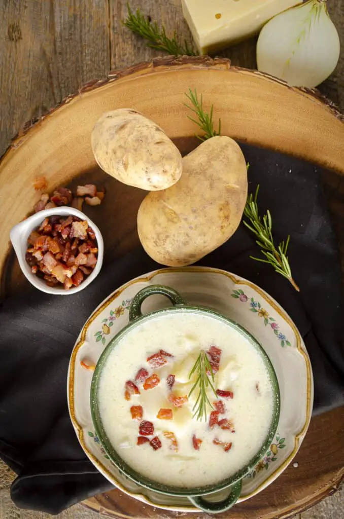 Looking down on a wooden table with a bowl of Gluten-free Swiss Potato Soup next to some russet potatoes, a tiny bowl of chopped bacon, and other ingredients.