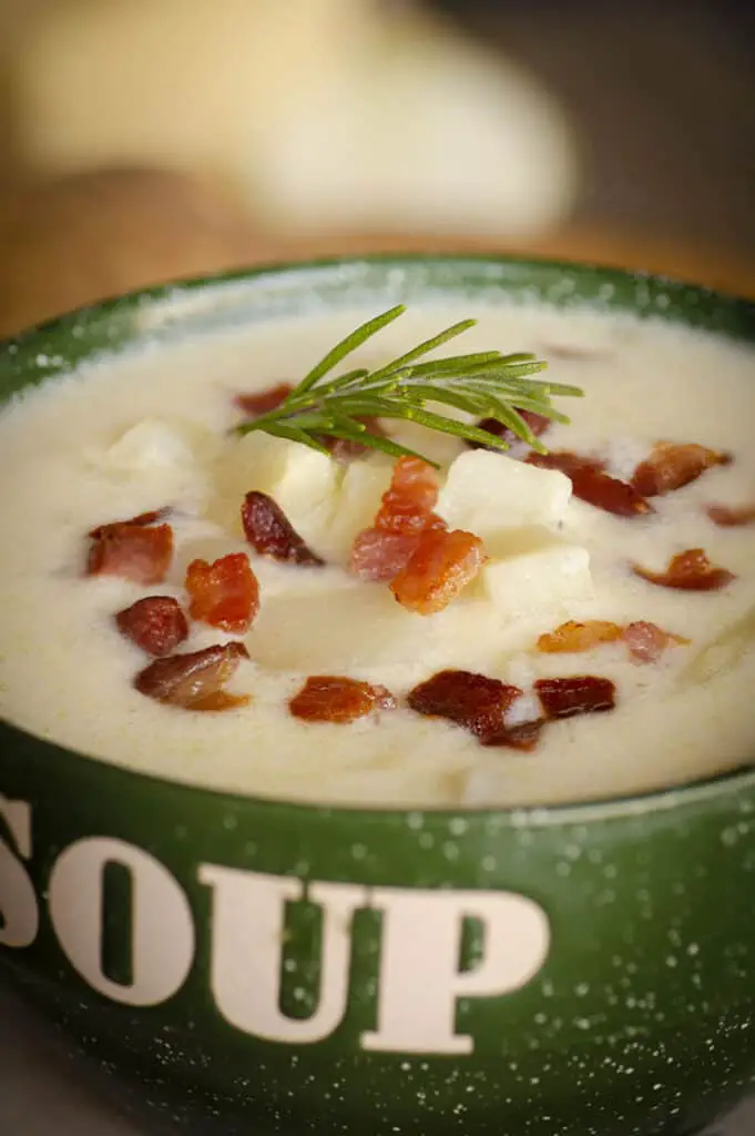 An extreme closeup of Gluten-free Swiss Potato Soup  in a green soup bowl with bacon bits and a rosemary sprig on top.