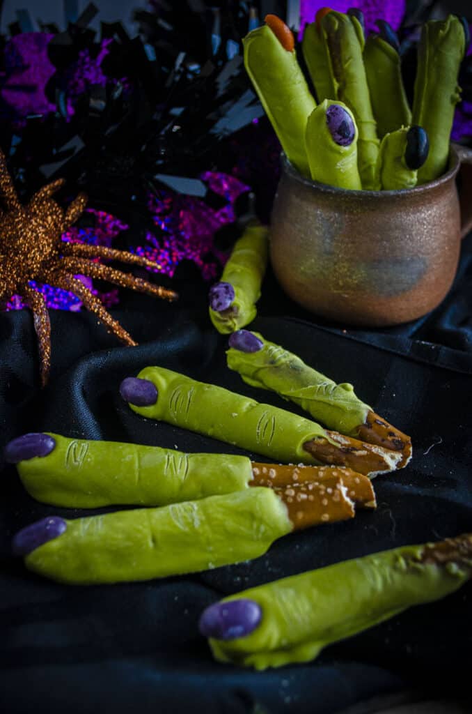 Green Witch Fingers Halloween Treats with purple jelly bean finger nails are displayed on a black cloth and in a small earthenware pot.