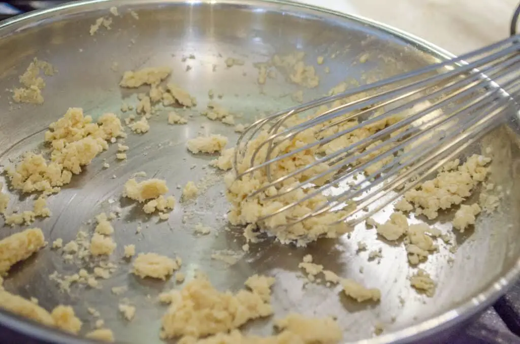 A whisk stirs clumps of butter and flour together in a saute pan.