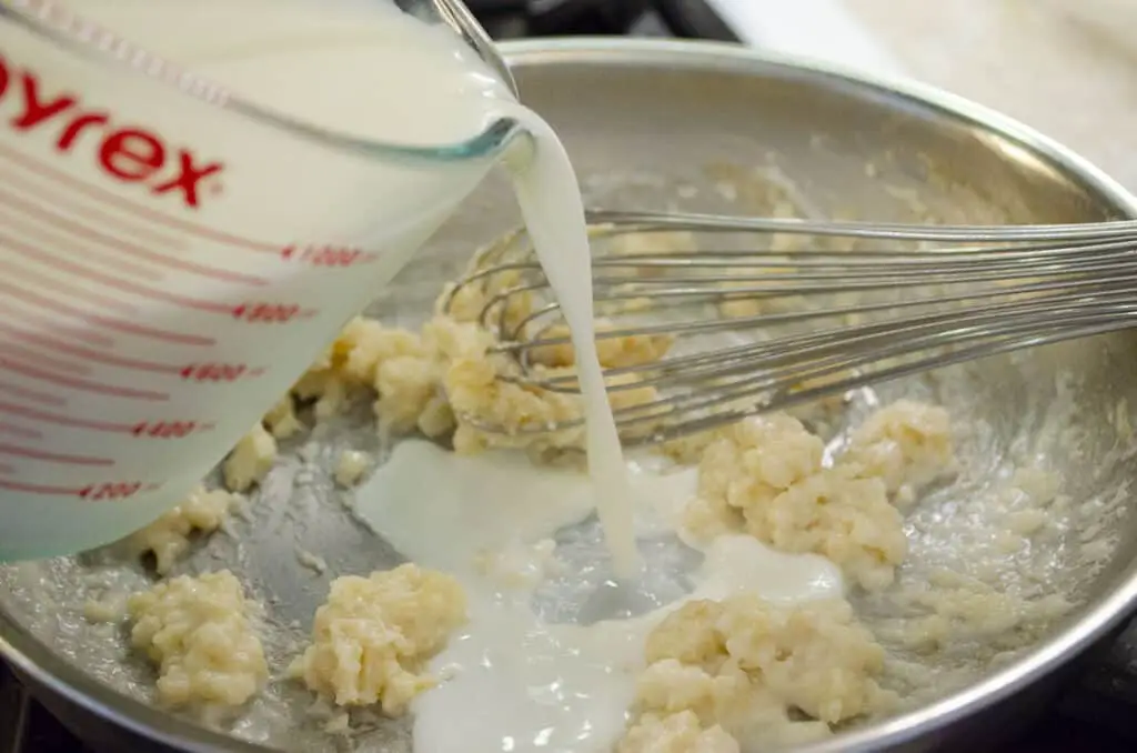 Milk is poured from a measuring cup into a stainless steel pan with clumps of flour and butter.