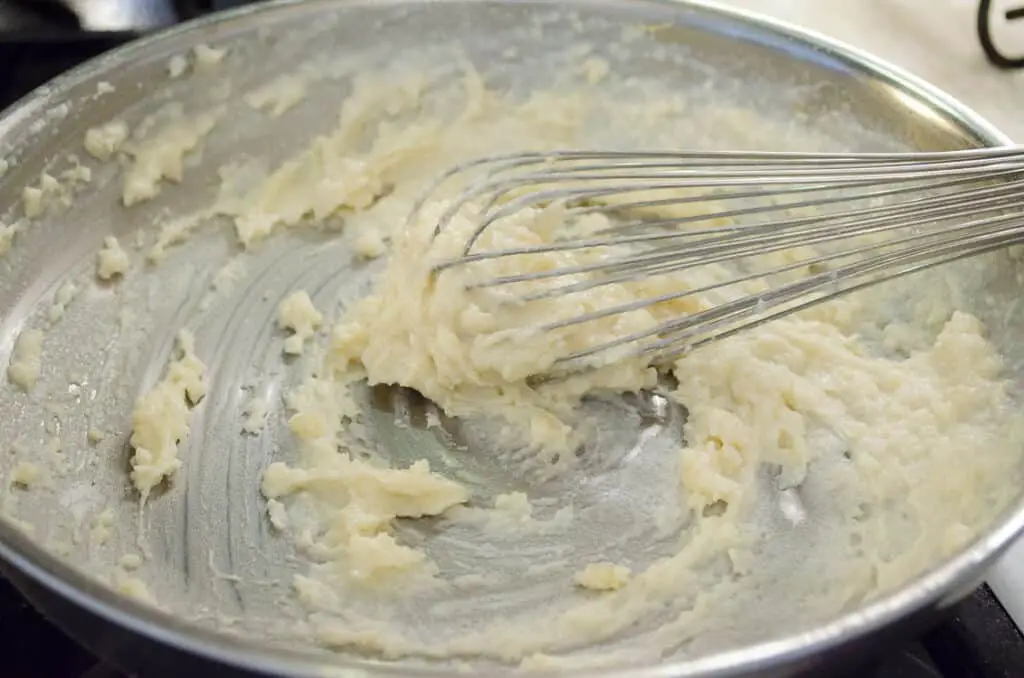 A thick paste of flour, butter and milk is stirred with a whisk in a stainless steel saute pan.