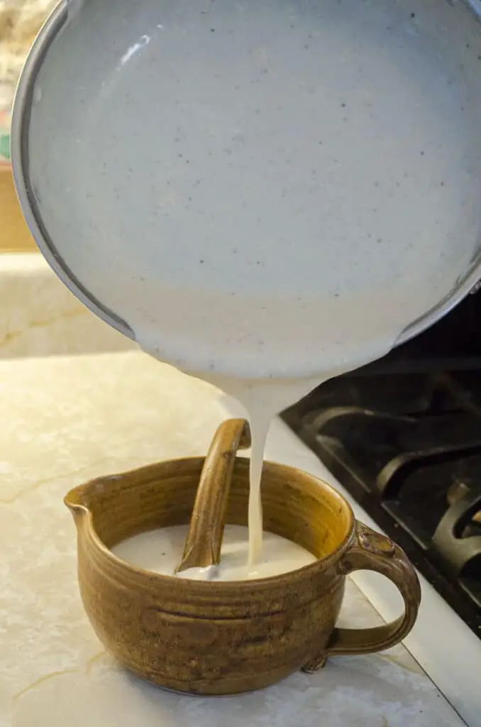 Homemade White Country Gravy is poured from a saute pan into a brown ceramic gravy boat with a ladle.