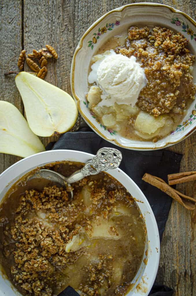 Looking down on a freshly baked Ginger-Pear Crisp baked in a white round ceramic dish next to a serving of Ginger-Pear Crisp on a plate with a scoop of vanilla ice cream.