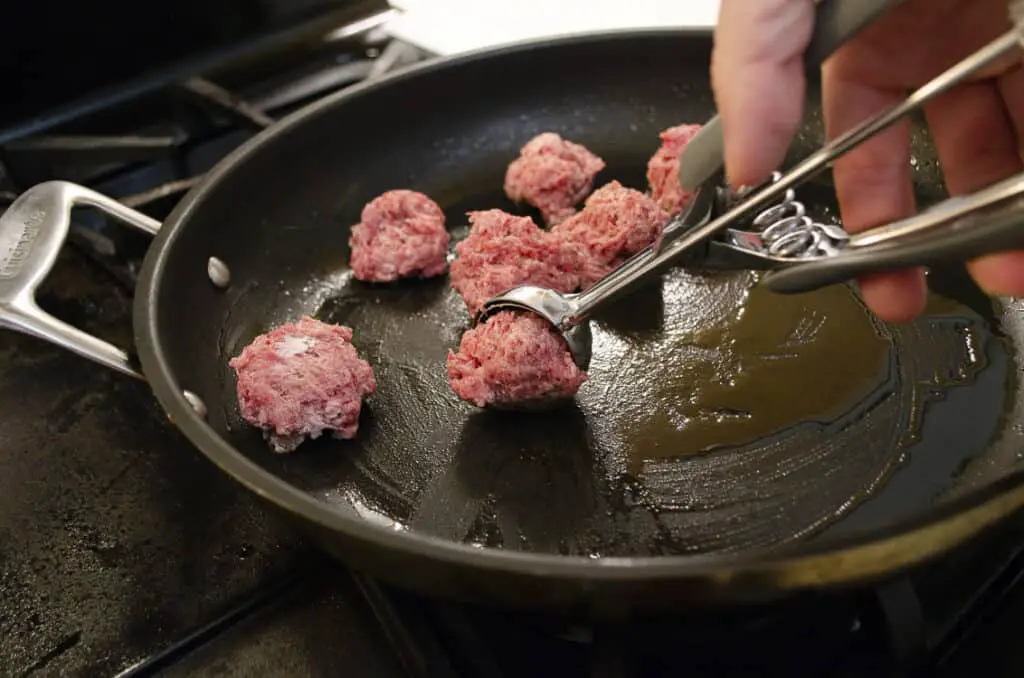 Raw meatballs being placed into a skillet with a small ice cream scoop.