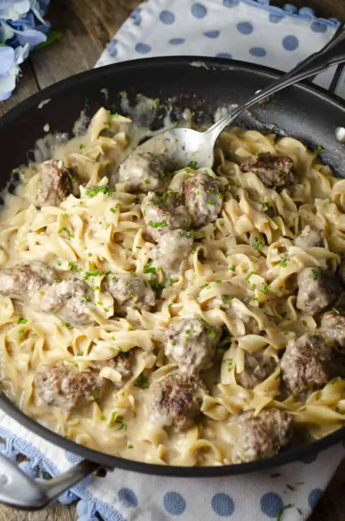 A skillet full of One-Pot Meatball Stroganoff sits on a wooden table with a large decorative serving spoon in it.