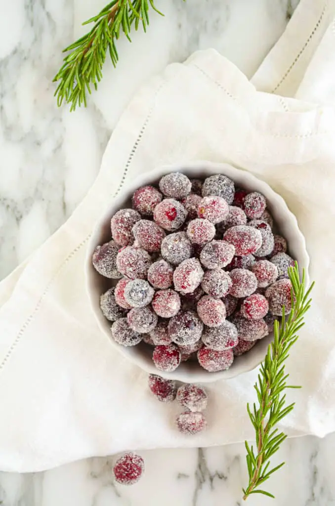 Looking down on Sparkling Sugared Cranberries in a white ceramic bowl sitting on a white napkin and white marble counter.