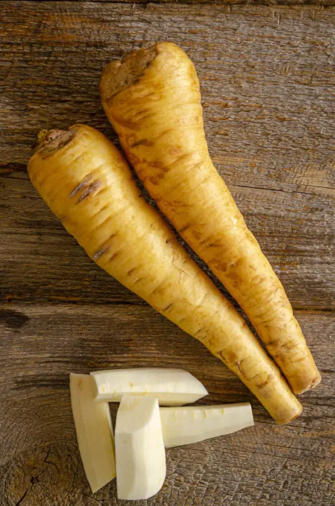 Two golden parsnips sit on a barn wood table next to some peeled and chopped pieces of fresh parsnip.