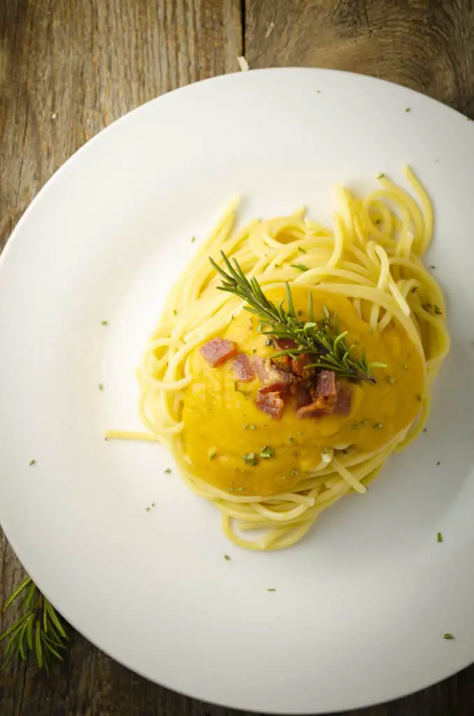 Looking down on a white plate with a serving of yellow Winter Squash Pasta Sauce over spaghetti garnished with bacon bits and a rosemary sprig.