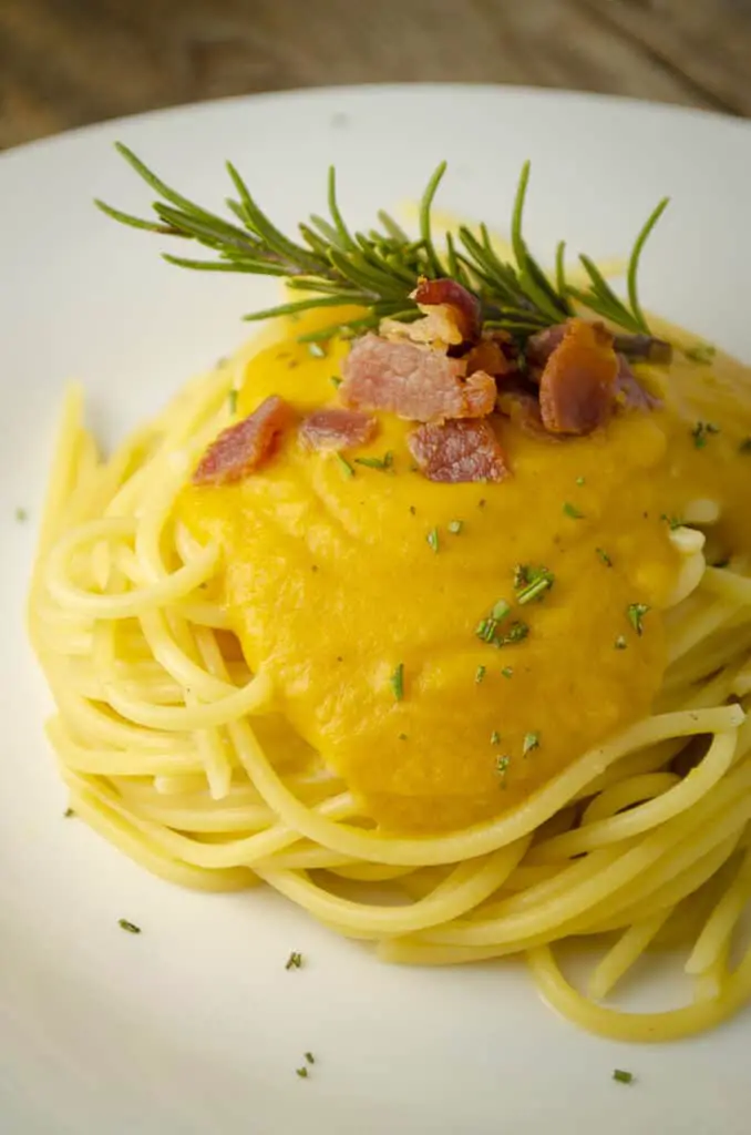 A closeup picture of a bed of spaghetti smothered in Winter Squash Pasta Sauce garnished with crumbled bacon, chives and a sprig of rosemary.