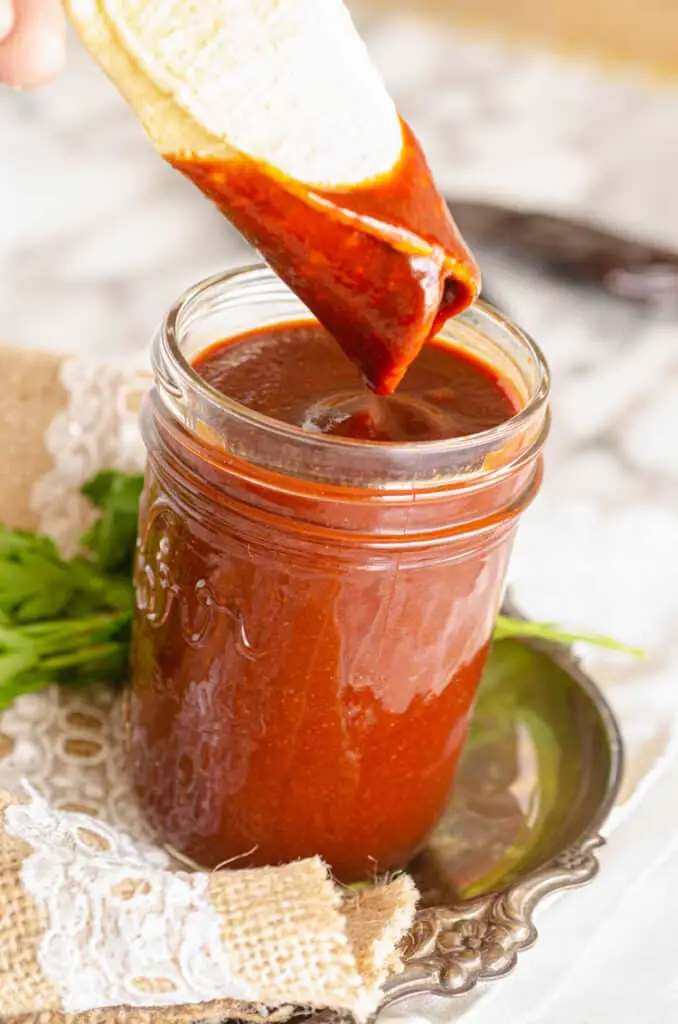 A closup picture of a mason jar filled with red chile. A rolled corn tortilla is held just above and has just been dipped into the chile sauce.