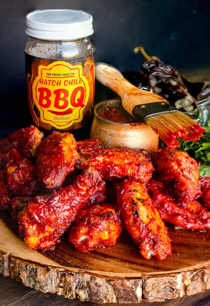 Hatch Chile Barbecue wings, sweet and spicy medium. Chicken wings covered in red sauce with a basting brush and a jar of barbecue sauce. All sitting on a wooden board.