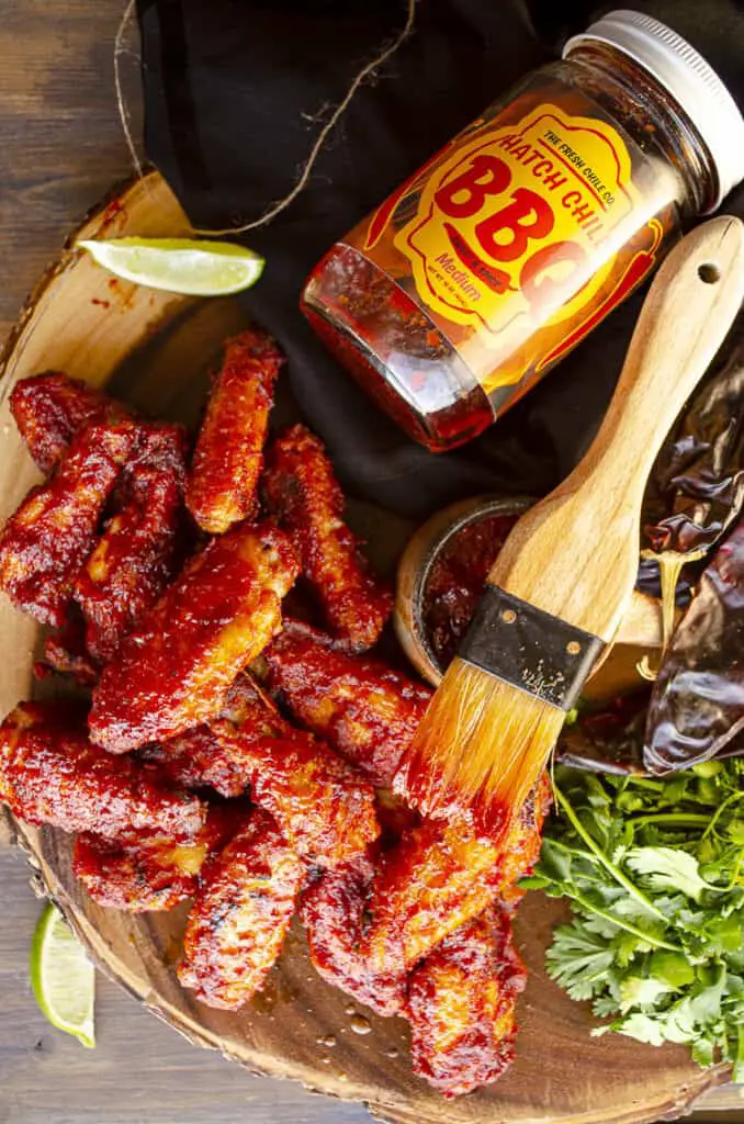 Looking down on a wooden tabletop showing Hatch Chile barbecue chicken wings with the jar of sauce, a basting brush, a gathering of fresh red cilantro, and dried red chile pods. 