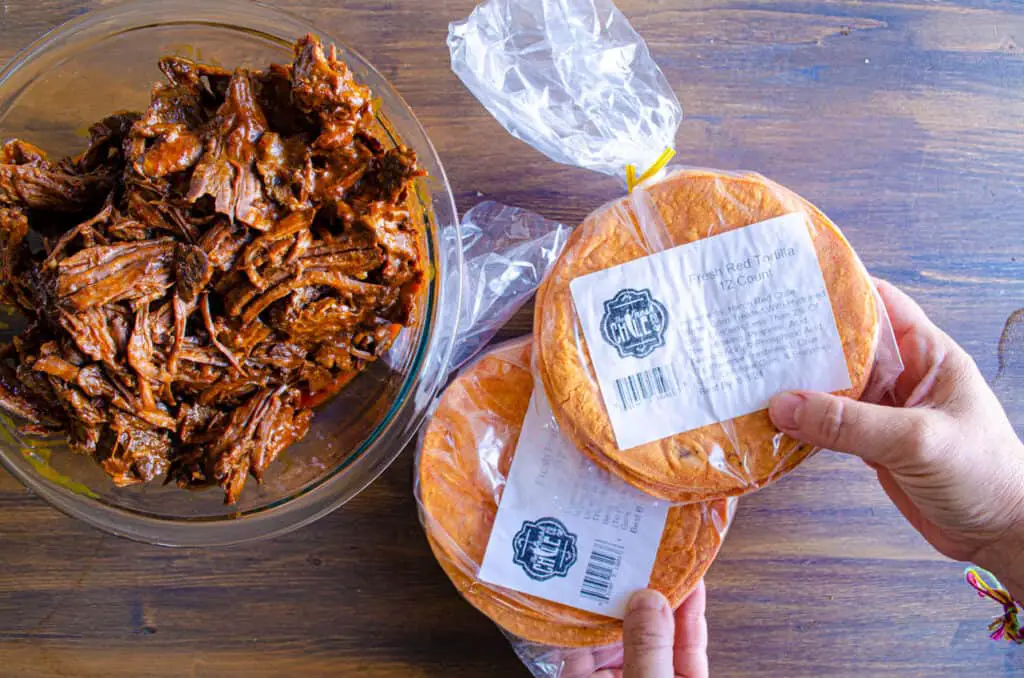 A person holds two packages of special red chile corn tortillas next to a bowl of shredded Birria beef.