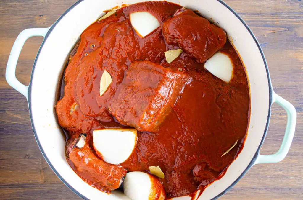 Looking down into a pot filled with beef chunks smothered in red chile sauce. Large chunks of onion and small bay leaves are scattered around on top.