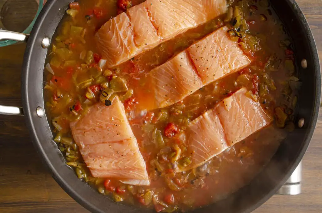 Looking down into a large non-stick skillet filled with salsa and raw, pink, salmon fillets placed skin side down (skin not visible).