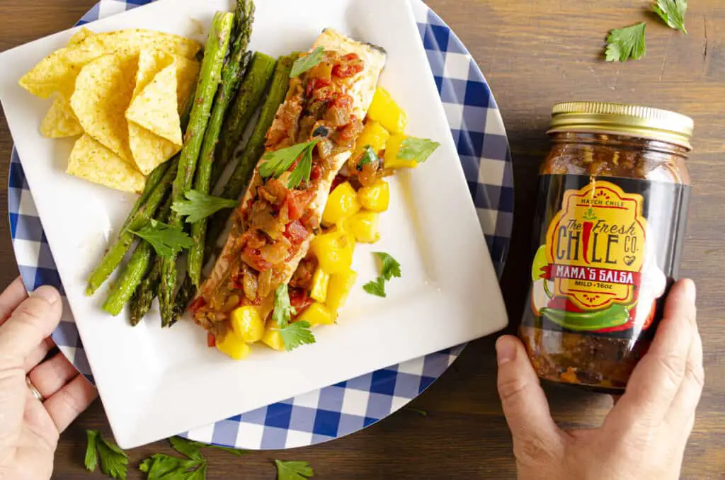 Looking down on a white square plate on a wooden table top displaying a serving of Easy, One-Pan Salsa Poached Salmon with Mango with grilled asparagus, mango chunks and tortilla chips. A person holds a jar of The Fresh Chile Co.'s Mama's Salsa next to the plate.