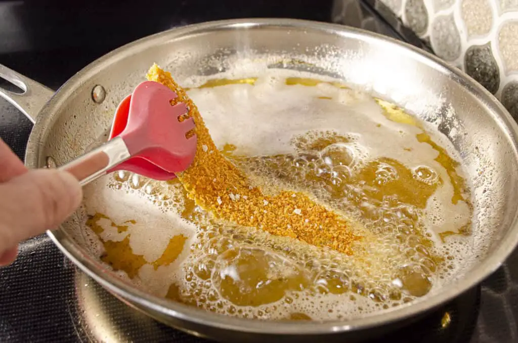 A breaded Hatch Green Chile is lowered into hot bubbling oil contained in a stainless steel frying pan.