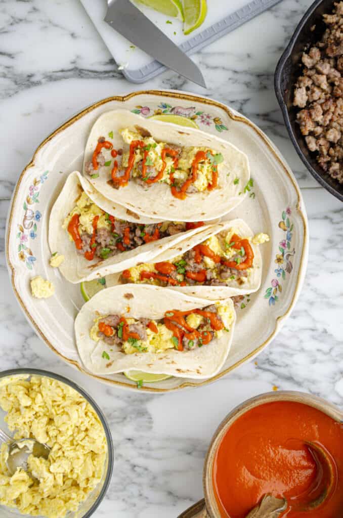 Looking down onto a white marble tabletop display of a plate of soft Fiesta Breakfast Tacos with Sausage & Red Chile Sauce, surrounded by bowls of red chile, scrambled eggs and crumbled sausage.