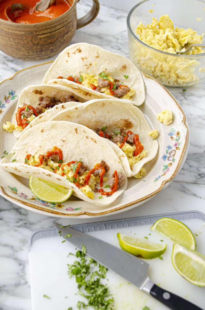 Fiesta Breakfast Tacos with Sausage & Red Chile Sauce being assembled onto a large plate surrounded by scrambled eggs, red chile sauce, and fresh lime wedges.