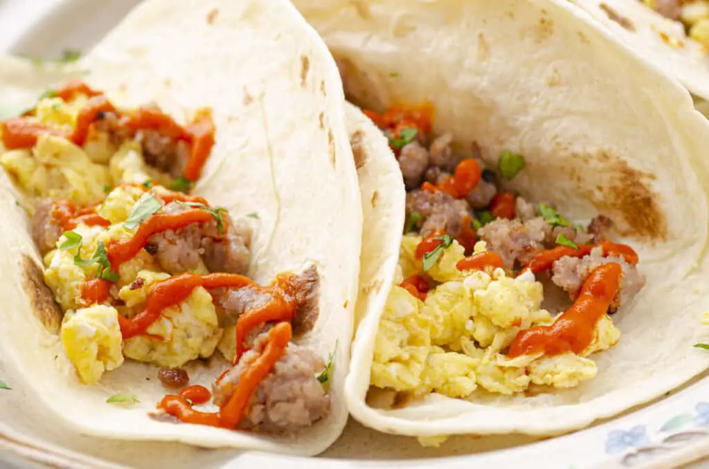 Looking almost horizontally and very closely at two soft Fiesta Breakfast Tacos with Sausage & Red Chile Sauce showing in detail the scrambled eggs and ground sausage, with a little drizzle of red chile sauce and chopped cilantro over the top.