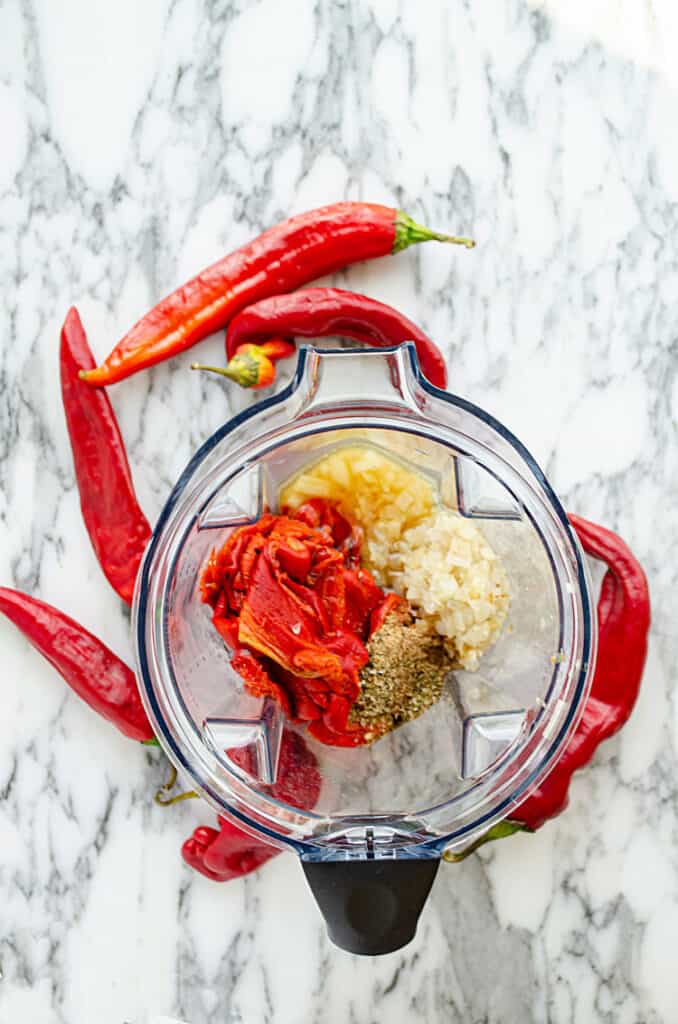 Looking down into the pitcher of a blender containing the ingredients to make red chile sauce from fresh red chile peppers. The pitcher is surrounded by whole red chile peppers.