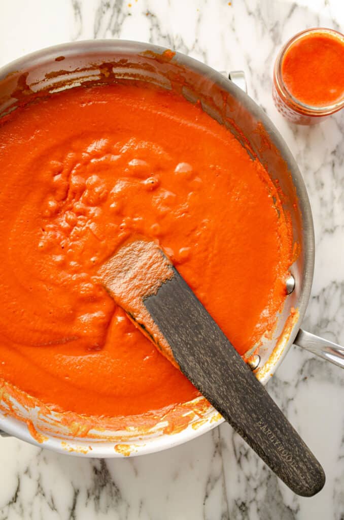 Looking down on a stainless steel skillet filled with fresh red chile sauce that is bubbling in the center. A wooden stir stick sits in the sauce and a mason jar of sauce sits at the side of the skillet.