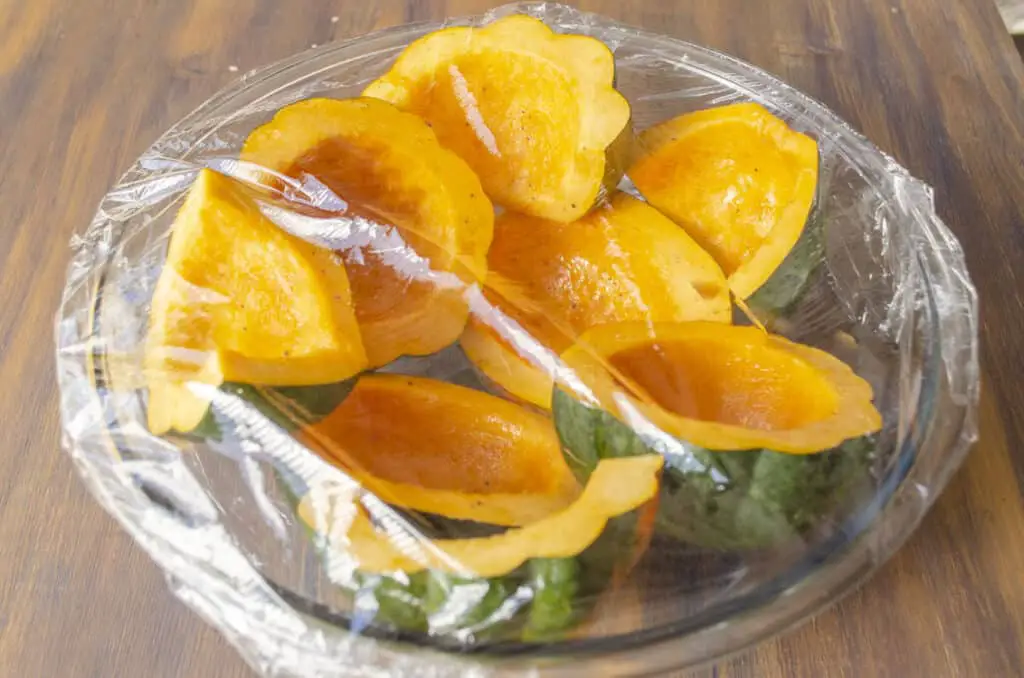 A large clear glass bowl containing cut halves of acorn squash, seeds removed. The bowl is covered with plastic wrap.