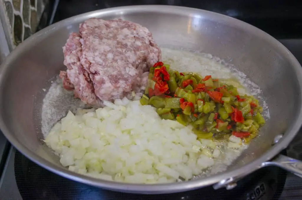 An aluminum skillet contains raw pork sausage, diced onion and Hatch green chile for making Green chile and Sausage stuffed Acorn Squash.