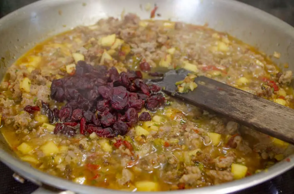 Cranberries and chicken broth are added to a pan sauteeing ingredients for Green chile and Sausage stuffed Acorn squash.