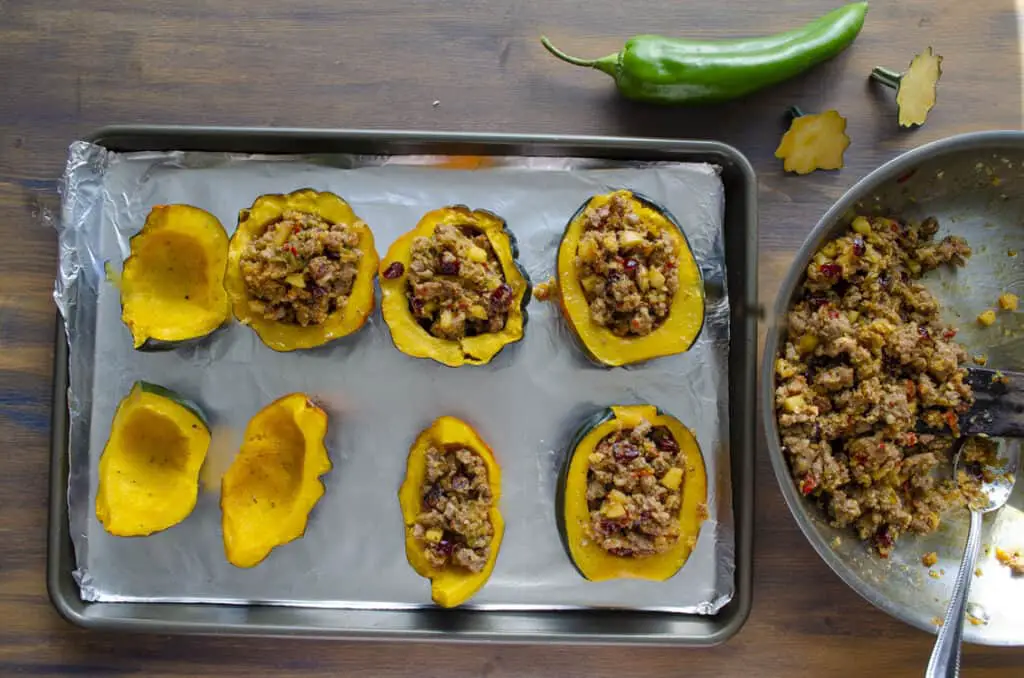 A rimmed baking sheet sits on a wooden table. On the sheet are several halved acorn squashes being filled with stuffing to make Green Chile and Sausage stuffed Acorn squash.