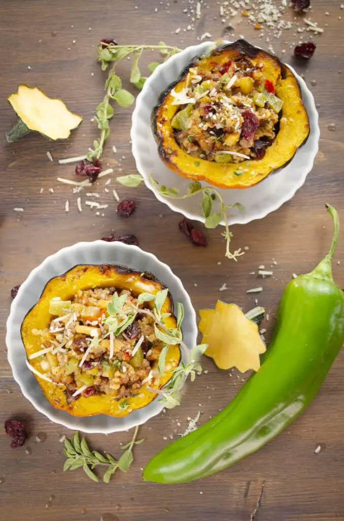 Looking down on two halves of an acorn squash filled with sausage and green chile stuffing. A whole green chile, sprigs of oregano, two acorn squash stems, shredded parmesan and dried cranberries are sprinkled around the stuffed squash.