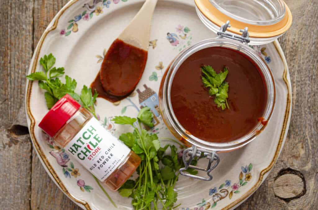 Looking down on a plate with an open jar filled with red chile sauce garnished with a sprig of cilantro. Next to the jar is a wooden spoon holding more chile sauce, and a Hatch chile store container of mild red chile powder. 