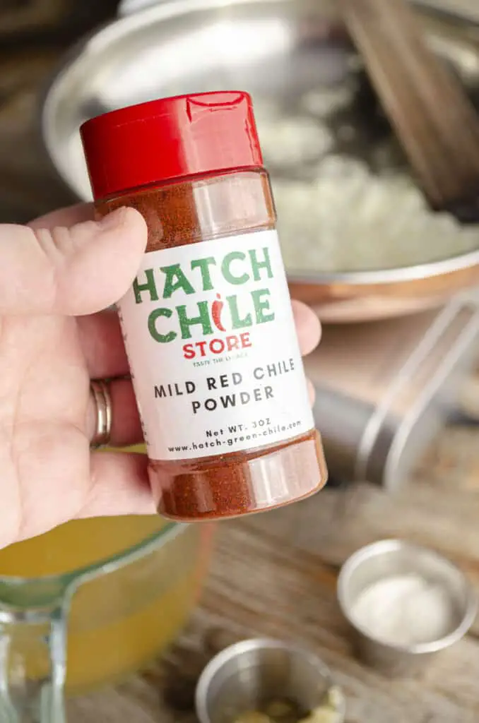 A closeup of a container of Mild Hatch Red Chile powder is being held by hand in front of a cooking station preparing to make Hatch Red Chile Sauce.